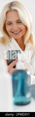 Vertical shot of beautiful Caucasian mature lady tapping on smartphone in cosmetology center Stock Photo