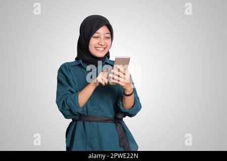 Portrait of excited Asian muslim woman with hijab holding and touching mobile phone. Advertising concept. Isolated image on white background Stock Photo