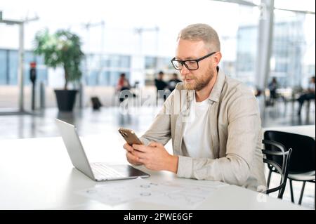 Focused successful caucasian man with a glasses and in casual stylish clothes, IT specialist, developer, programmer, sit at a work desk, using smart phone, chatting with the client, responds to email Stock Photo