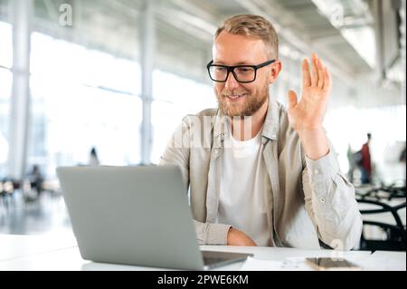 Online video conference. Positive friendly caucasian man, seo, mentor, talking on video call with colleagues or students sitting at the table in the office, having a brainstorm, greeting them, smiling Stock Photo