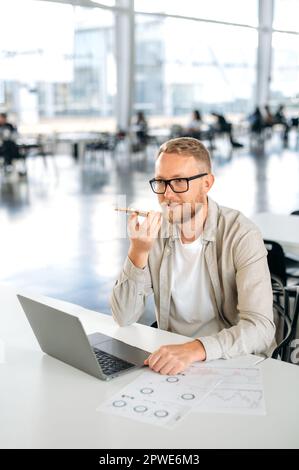 Vertical photo of positive successful caucasian man with a glasses, IT specialist, developer, programmer, sit at a work desk, using smart phone, talking on speakerphone, recording a voice message Stock Photo
