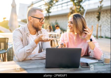 Positive successful Caucasian colleagues, a man and a woman, business partners, sit outdoors in cafe with a laptop, discuss work plans and prospects, share ideas, smile. Partnership, cooperation Stock Photo