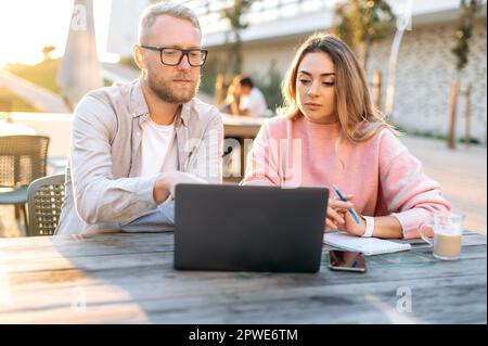 Focused partners, a man and a woman of Caucasian ethnicity, are sitting in a cafe at a table with a laptop, discussing the terms of cooperation, planning a startup, promotion and strategy Stock Photo