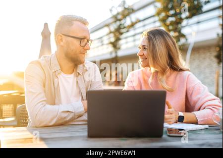 Partnership, cooperation. Two successful Caucasian colleagues, a man and a woman, business partners, sit outdoors in cafe with a laptop, discuss work plans and prospects, share ideas, smile Stock Photo