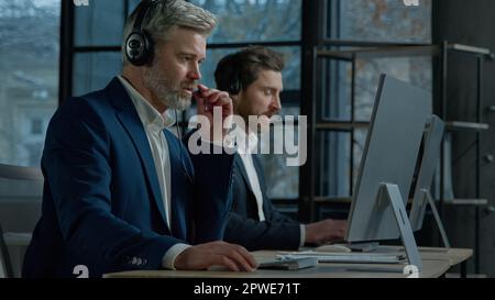 Two men call center service operators work in office talk to client using headset on computer concentrated sales agents sell online answering incoming Stock Photo