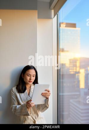 Young Asian business woman using digital tablet standing at office window. Stock Photo