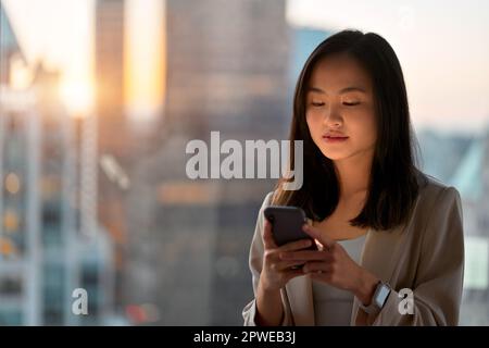Young Asian professional business woman using phone standing at office window. Stock Photo
