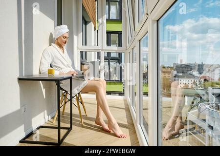 Barefoot woman in a bathrobe sits on balcony of her new high-rise apartment and using laptop Stock Photo