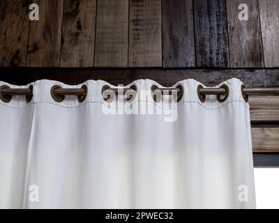 Close-up of white wall curtain on wooden curtain rail bar decorated on the wood plank wall of tropical building over the glass door or window inside r Stock Photo