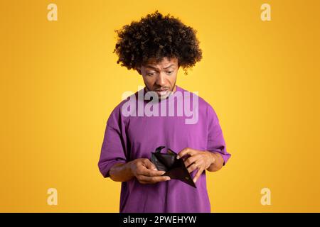 Sad shocked adult african american curly man in purple t-shirt looks at empty wallet Stock Photo