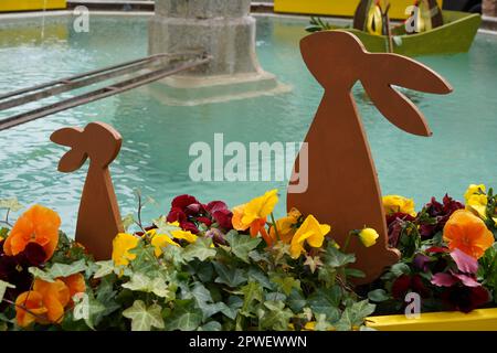 Two outdoor Easter bunnies, big and small, made of wood and placed among flowers. There is fountain water on the background. Stock Photo