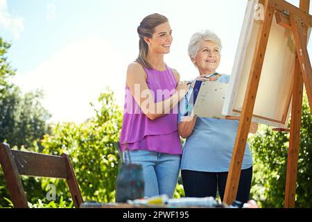 She loves watching her mother paint. a senior woman and her adult daughter painting in a park. Stock Photo