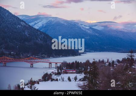 Winter view from Gyro Park of the big orange bridge across the west arm of Kootenay Lake and the snowcapped peak of Kokanee Glacier mountain catching Stock Photo