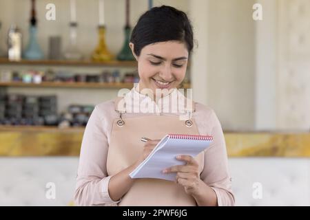Pretty Indian waitress writing in notepad standing at workplace Stock Photo