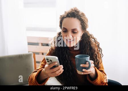 Happy young woman enjoying mobile technology while drinking coffee at home. Smiling lady using smartphone for online shopping and social media at home Stock Photo