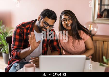 Happy indian family couple cuddle at desk make video call to friends using laptop webcam. Loving young spouses look at computer screen waving hands in Stock Photo