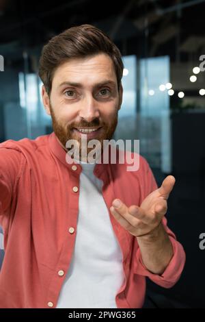 Vertical photo. Close-up photo. Young man freelancer, blogger in red shirt greets and talks to the camera, smiles, waves, records video, blog. Stock Photo