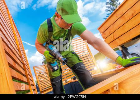 Caucasian Worker in His 40s Making Holes on the Bottom of Wooden Planters Using Hole Saw Bit and Drill Driver. Garden Small Architecture. Stock Photo