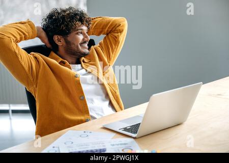 Satisfied indian or arabian man, company employee, sits at his work desk, rests from work in a laptop, relaxes with his hands behind his head, dreaming about weekend, smiles. Relaxation and rest Stock Photo