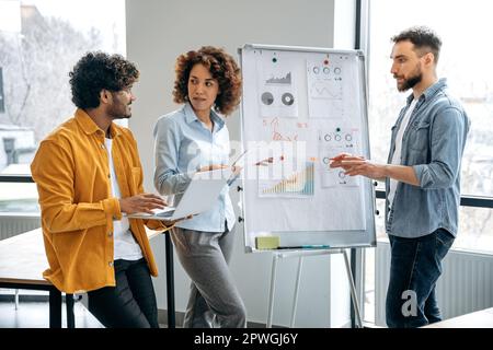 Group of a creative positive clever multiracial colleagues of financial department, stand near whiteboard in office, developing and planning strategy, thinking about ideas, analyze risks and profit Stock Photo