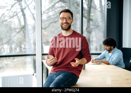 Excited successful caucasian man in casual wear with glasses, freelancer, creative manager, holds his smart phone while sitting on a table in the office, got good news, looks at camera, smiles happily Stock Photo