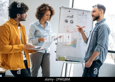 Brainstorming. Successful creative coworkers are working together on a new project, standing near white marker board with graphs in the office, predicting income, analyzing risks, setting goals Stock Photo