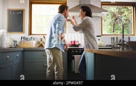 Tell me how it tastes...an affectionate young woman feeding her husband a spoonful of her food while cooking in their kitchen. Stock Photo