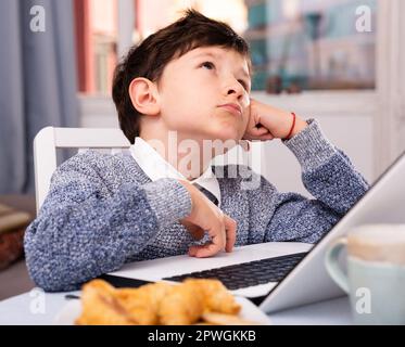 Unhappy preteen boy sitting near laptop at home table Stock Photo