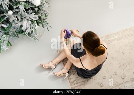 View from above at a woman in black glitter dress sitting on the floor near a Christmas tree and taking violet silk scarf out of a gift box Stock Photo