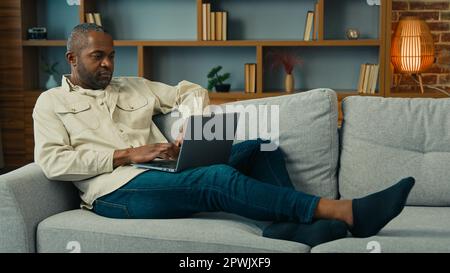 Mature african american man lying on sofa relax using computer businessman freelancer typing on laptop work internet at home ethnic male entrepreneur Stock Photo