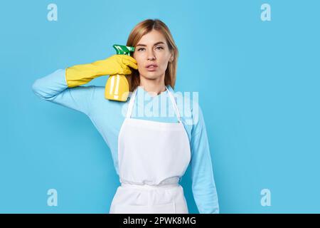 tired blonde woman in rubber gloves and cleaner apron holding detergent sprayer and pointing to head on blue background. Stock Photo