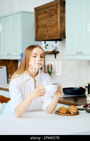 Pensive young woman sits behind the kitchen table with cup of coffee and thinking about something Stock Photo