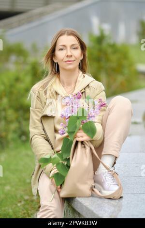 Pretty senior woman sits on embarkment with branch of lilac in her hands Stock Photo