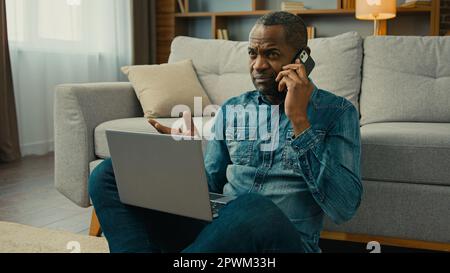 Mature african american man businessman freelancer talking on mobile phone discuss business deal using laptop at home in living room remote make order Stock Photo