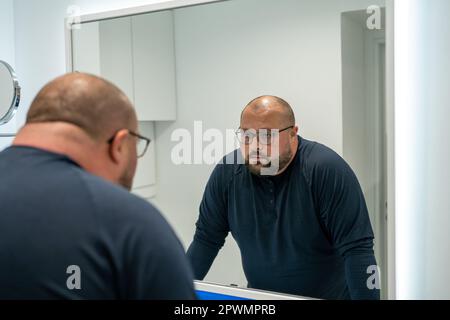 Confused upset overweight middle aged man looking at mirror in bathroom at home. Stock Photo