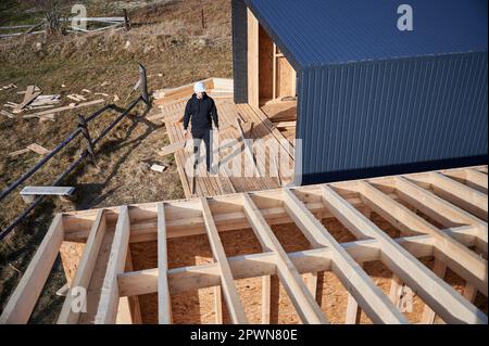 Male engineer building wooden frame house. Man developer on construction site, inspecting quality of work on sunny day, holding hammer in hands. Stock Photo