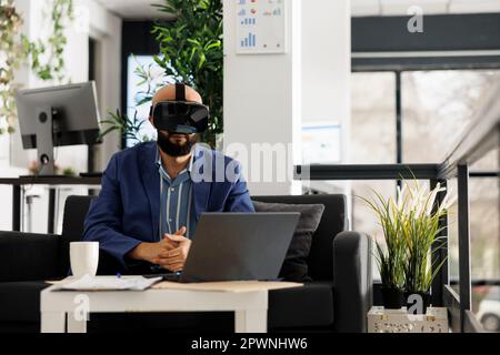 Arab business worker analyzing financial data in metaverse using vr glasses in start up office. Young man professional brainstorming wearing virtual reality headset in green open space Stock Photo