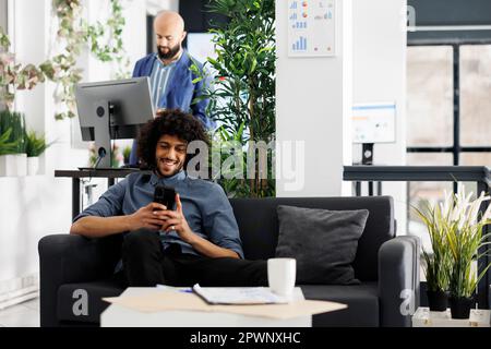 Cheerful arab entrepreneur chatting online on smartphone while sitting on couch in business start up office. Laughing businessman watching funny video on mobile phone in coworking space Stock Photo