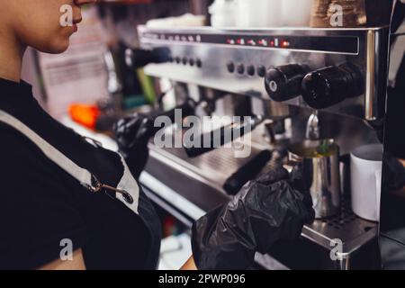 Barista in black gloves presses the button for pouring boiling water into metal cup while preparing coffee drink or cocktail. Close up of part of prof Stock Photo