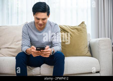 Asian older middle aged man relaxing sitting on sofa using smart mobile phone at home, Happy mature men smiling chatting with social media on smartpho Stock Photo