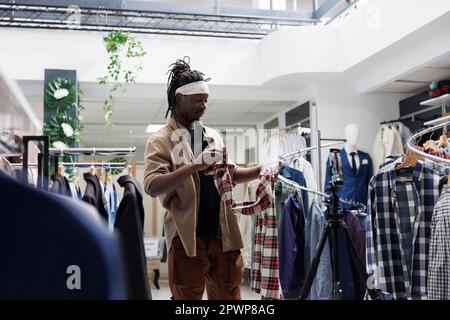 African american influencer speaking about clothing brand in store while recording video on smartphone. Social media fashion blogger showing shirt and giving review for online followers Stock Photo