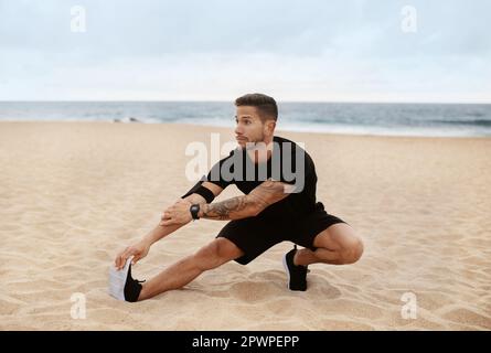 Young sporty man in sportswear doing leg stretching, preparing for exercising on ocean beach in the morning Stock Photo