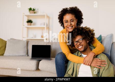 Black Couple Posing With Laptop, Embracing Smiling To Camera Indoors Stock Photo