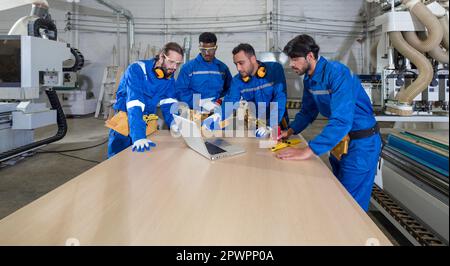 Group of multiethnic technicians in blue mechanic jumpsuits meeting,  using laptop computers on large wooden table. Production line atmosphere in furn Stock Photo