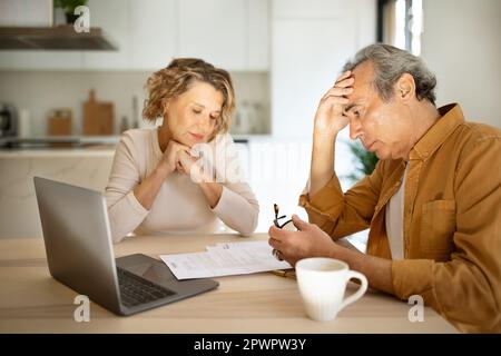 Stressed elderly couple checking financial papers, sitting with laptop and looking at loan documents, kitchen interior Stock Photo