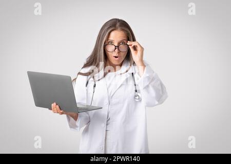 Surprised senior female doctor in white coat taking off glasses and looking at camera, using laptop pc, light background Stock Photo