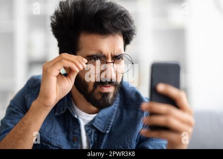 Eyesight Problems. Indian Man In Eyeglasses Looking At Smartphone Screen And Squinting Stock Photo