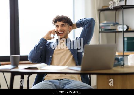 Cheerful Arabic Guy Talking On Cellphone Sitting At Workplace Indoors Stock Photo