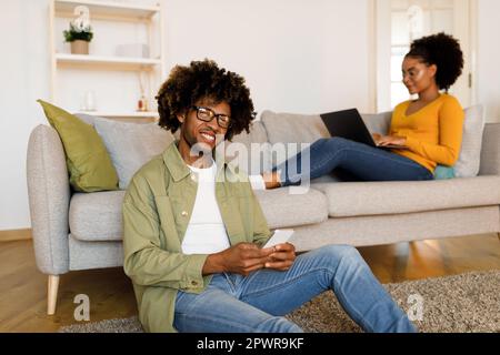 African American Man Holding Cellphone, Woman Using Laptop Working Indoors Stock Photo