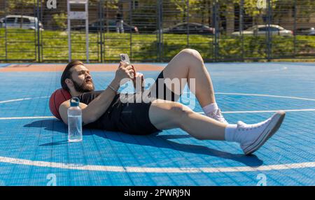 Portrait of tired athletic lebanese man in sportswear after playing basketball game. Guy lying down on ball relaxing use smartphone texting share messages on urban city court. Fitness on sports field Stock Photo